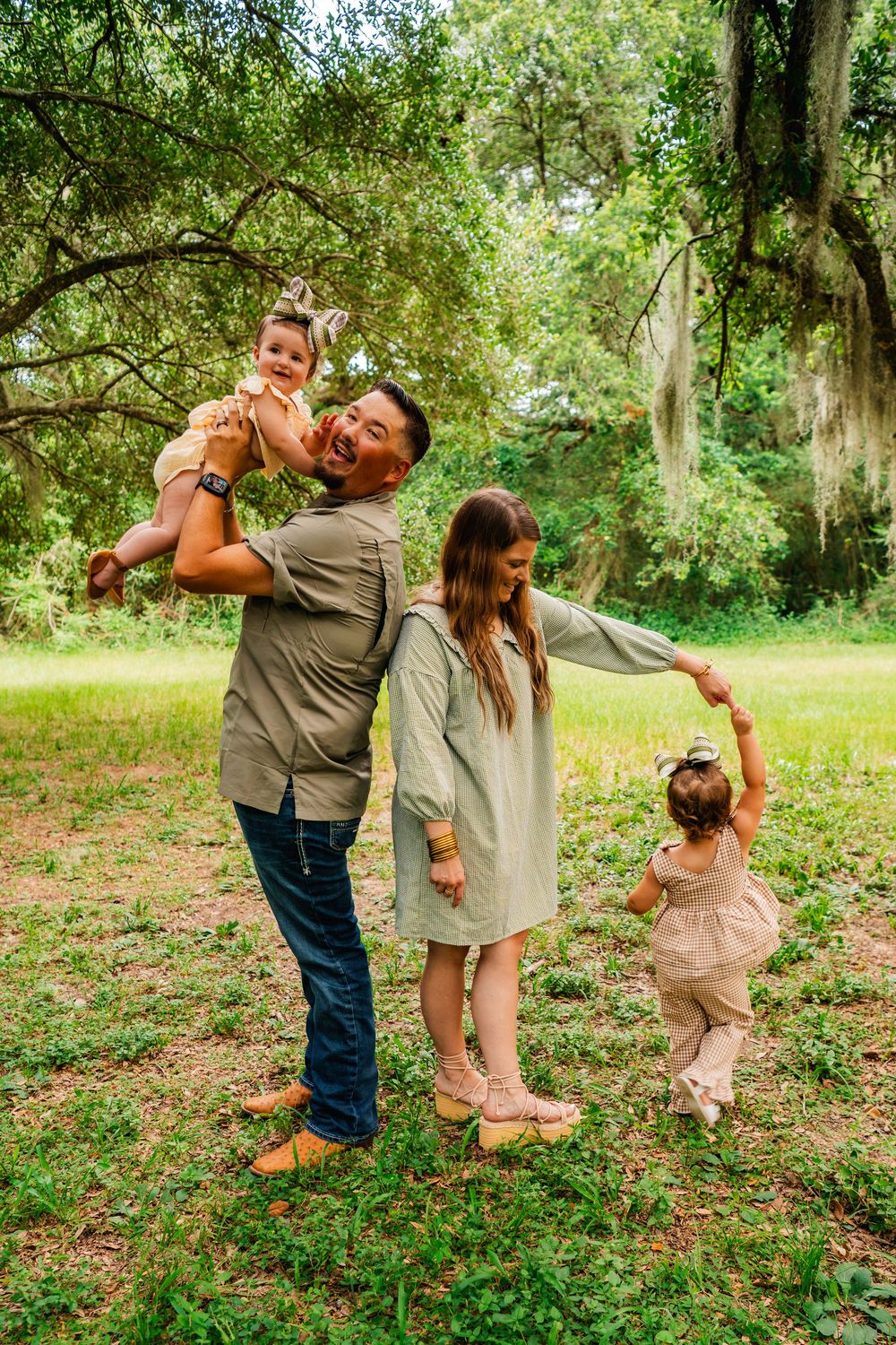 A joyful family of four plays outdoors under oak trees, with dad lifting a baby while mom twirls a toddler.