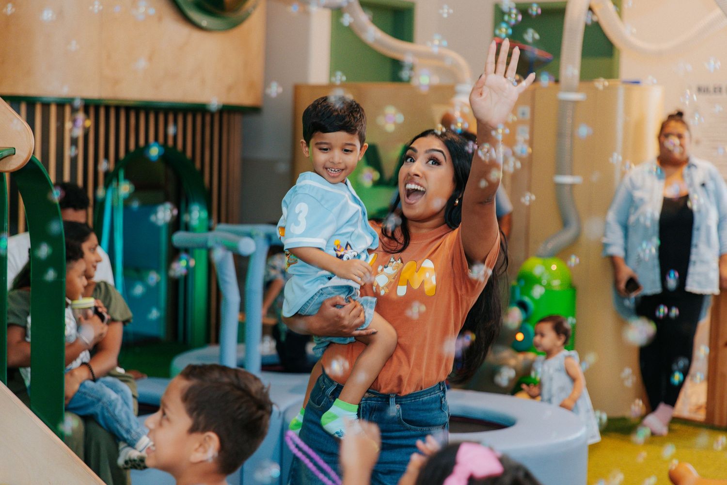 Woman holds toddler while reaching for bubbles in colorful indoor play area with children around.