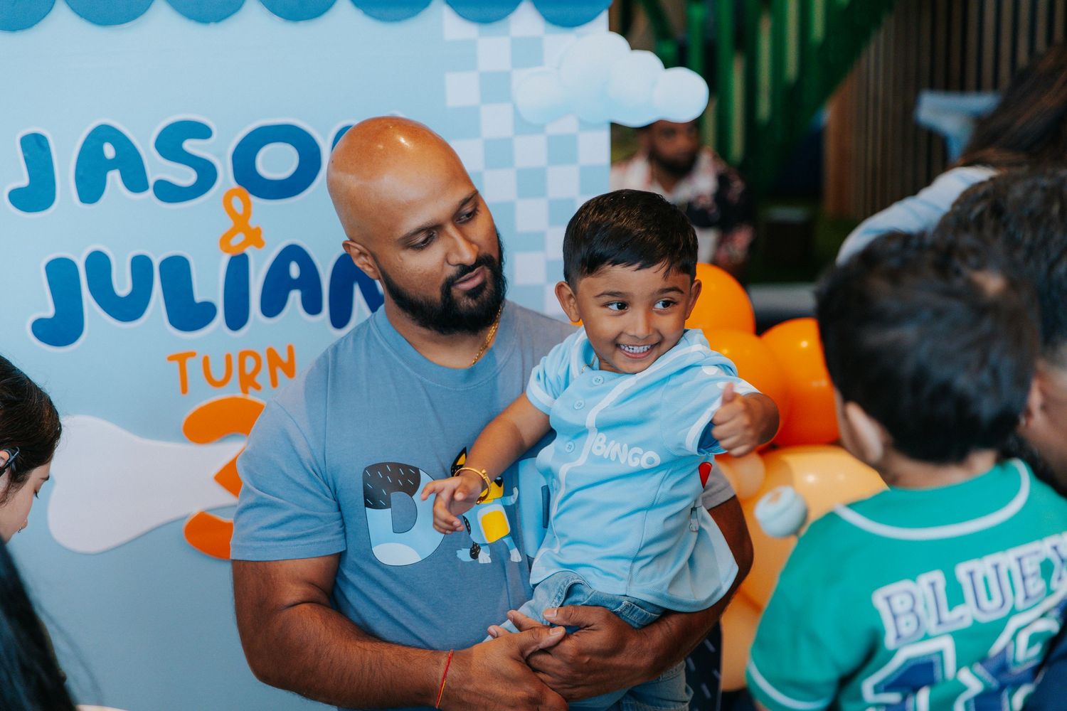 A man holds a toddler in a light blue outfit at Jason and Julian's birthday party with colorful balloon decorations.