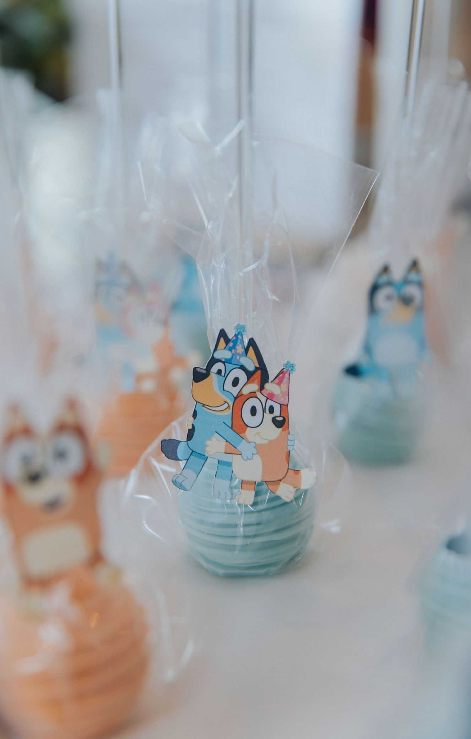 Bluey themed cake pops in pastel orange and blue, wrapped in clear bags with cartoon character toppers for a kids party.