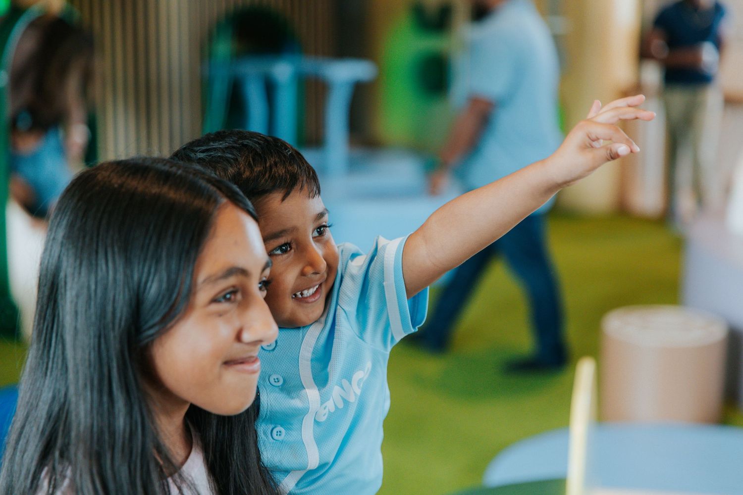 A young boy in a blue shirt points excitedly while a girl smiles beside him at a colorful indoor event.