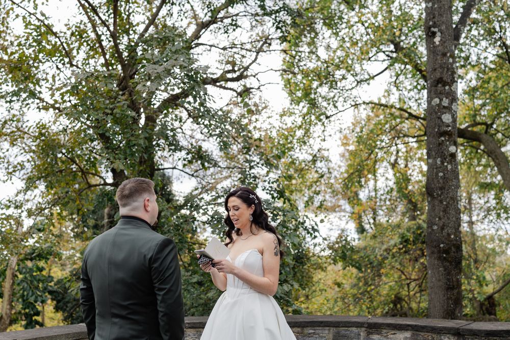 Bride reads vows to groom outdoors during intimate wedding ceremony surrounded by lush green trees.