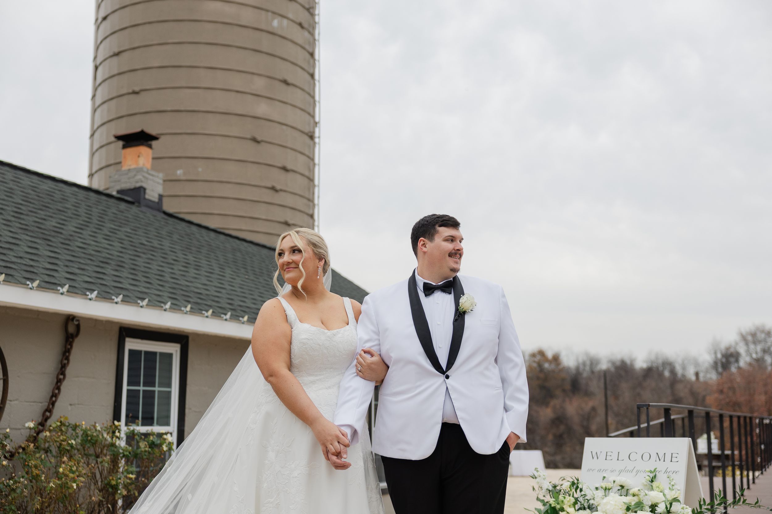 Bride and groom walking hand in hand outdoors near a rustic barn venue with a silo backdrop on their wedding day.