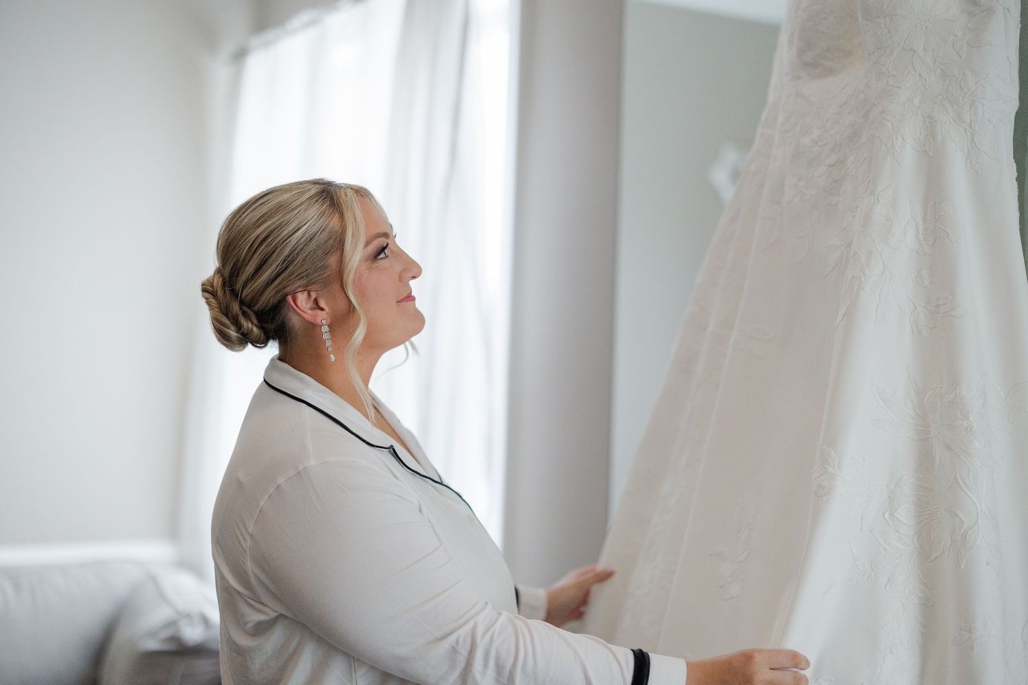 Bride in silk robe admiring her lace wedding dress hanging by a bright window before the ceremony.