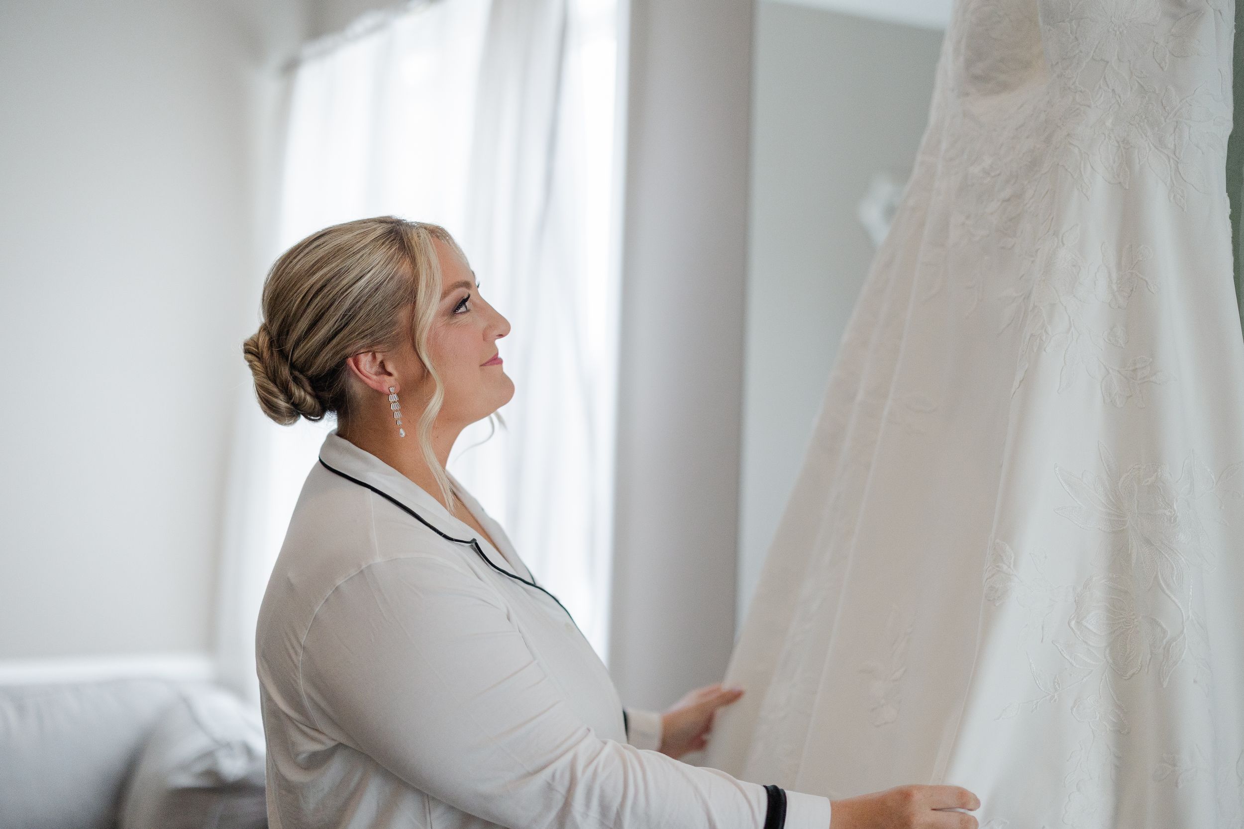 Bride in silk robe admiring her lace wedding dress hanging by a bright window before the ceremony.