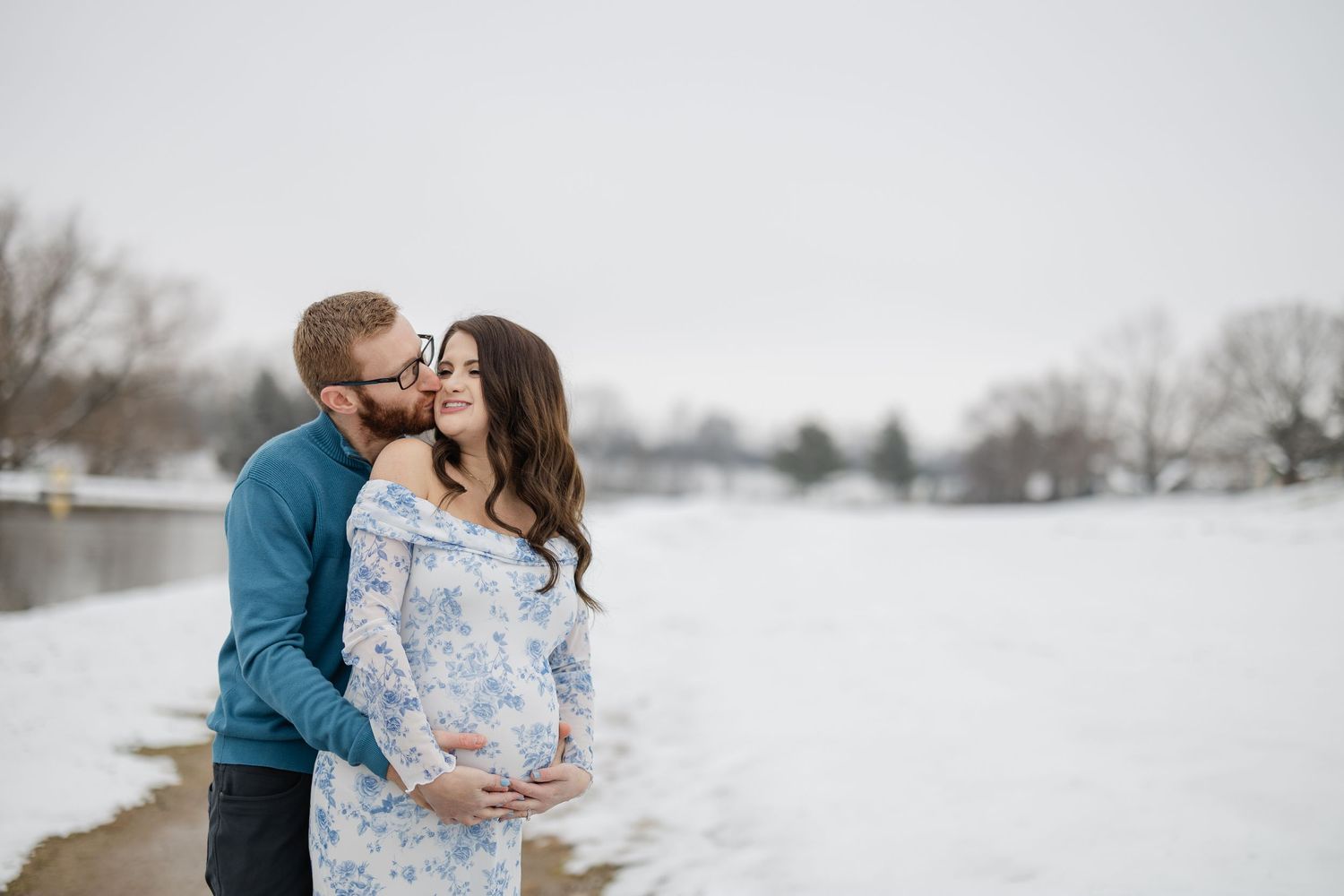 Couple sharing a tender moment in snowy winter landscape, pregnant woman in floral dress with partner kissing her cheek.