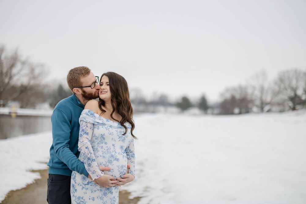 Couple sharing a tender moment in snowy winter landscape, pregnant woman in floral dress with partner kissing her cheek.