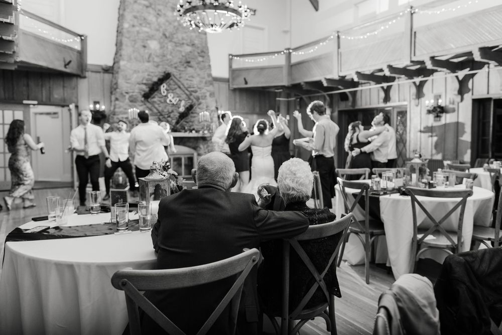 Black and white photo of wedding guests dancing and celebrating at a reception venue with round tables and ambient lighting.