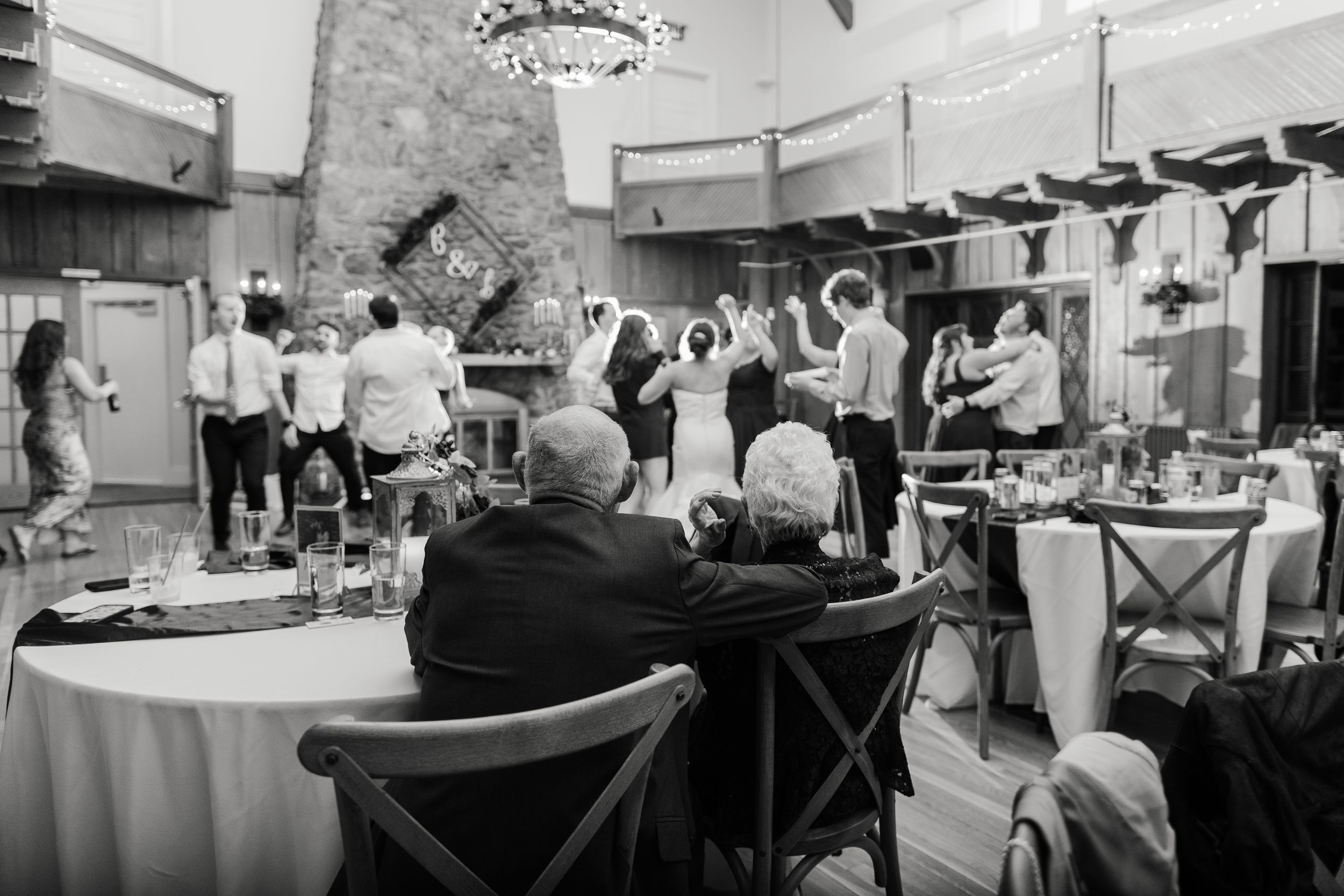 Black and white photo of wedding guests dancing and celebrating at a reception venue with round tables and ambient lighting.