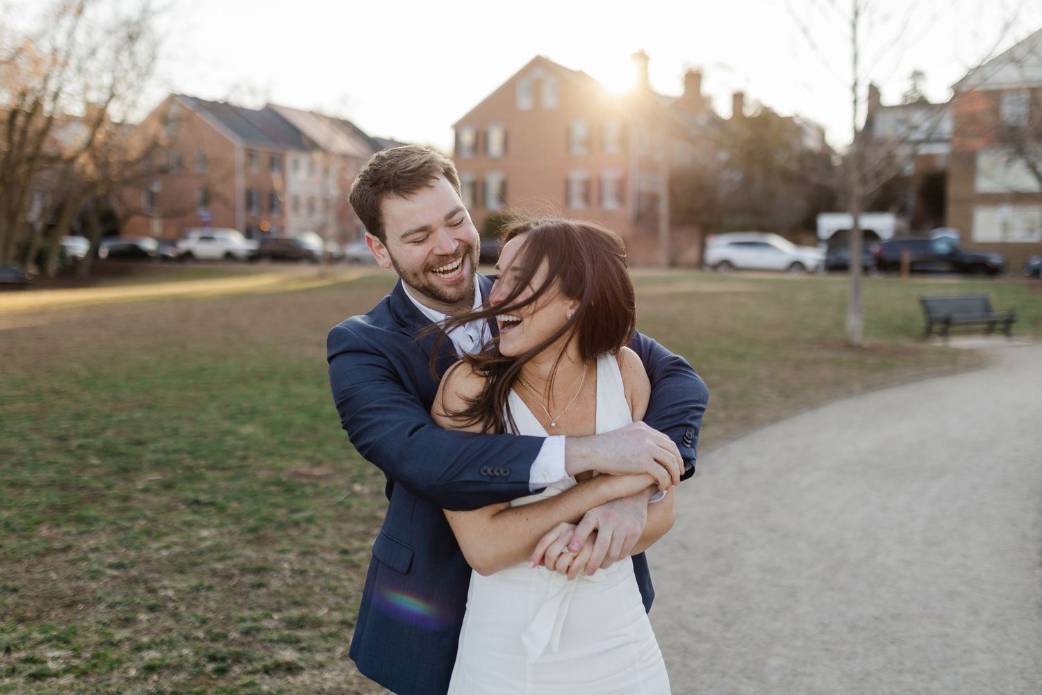 A happy couple embraces and laughs together on a path during golden hour with brick buildings in the background.