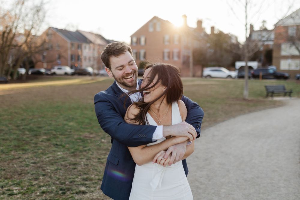 A happy couple embraces and laughs together on a path during golden hour with brick buildings in the background.