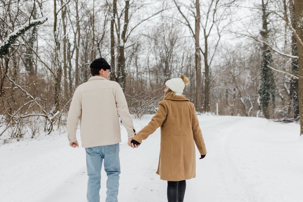 Two people holding hands while walking down a snow-covered path through bare winter trees in matching warm coats.
