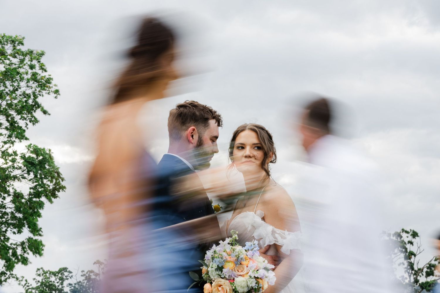 Artistic wedding photo of couple on overcast day holding floral bouquet with motion blurs of guests in foreground.
