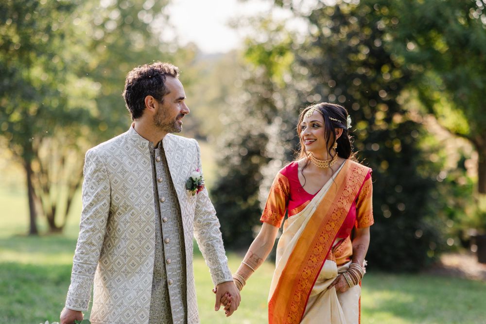A couple in traditional Indian wedding attire walking hand in hand through a sunlit garden setting.
