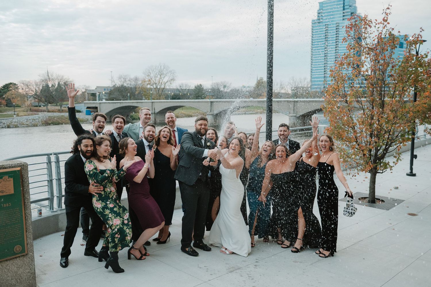 Wedding party celebrating on riverside with city skyline in background.