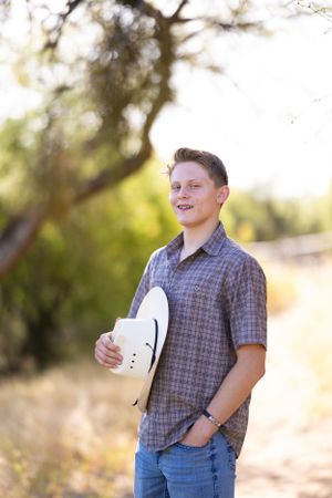 Person stands outdoors in plaid shirt holding white hat against natural desert background.