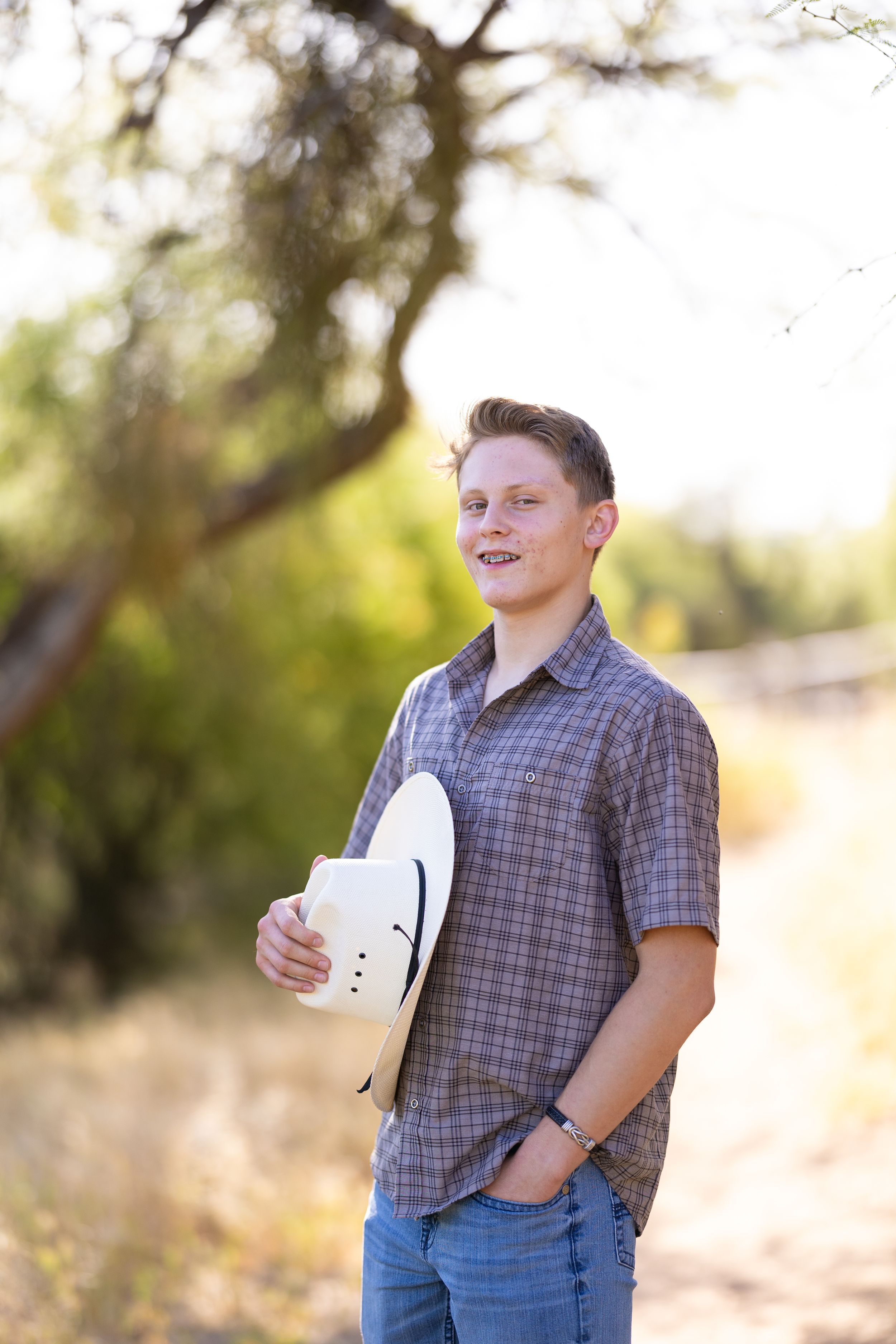 Person stands outdoors in plaid shirt holding white hat against natural desert background.
