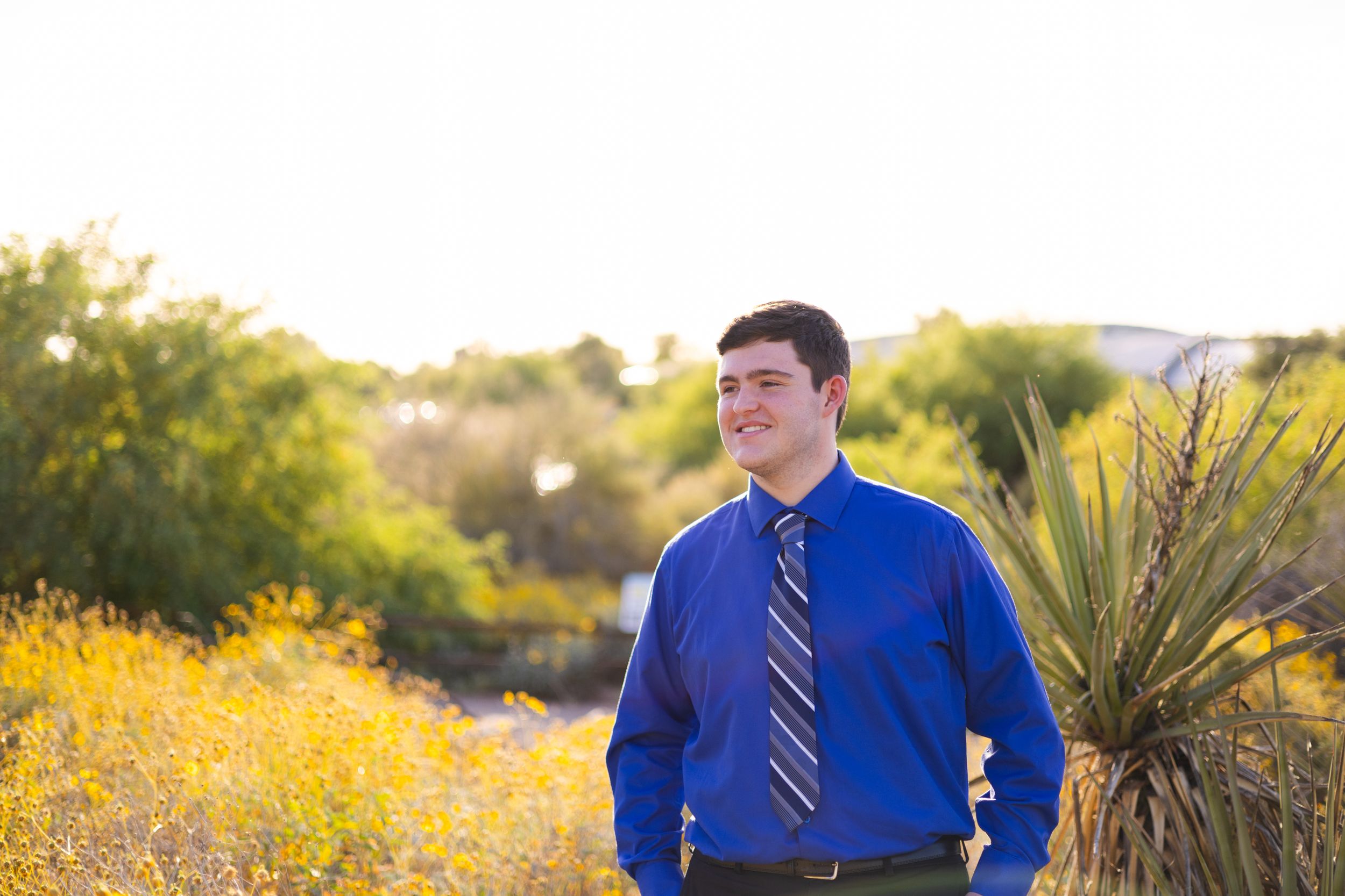 Person in blue dress shirt and tie poses outdoors with yellow wildflowers in background.
