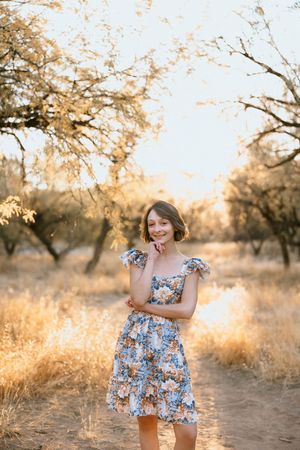Young woman in floral dress smiling outdoors among golden trees at sunset.