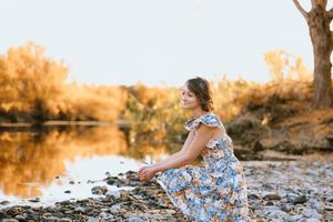 Young woman in floral dress sitting on rocky riverbank during golden hour sunset with autumn trees reflected in water.