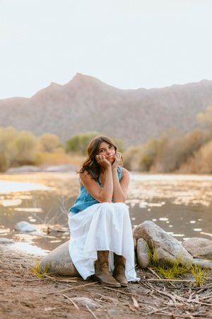 Woman in blue top and white skirt sitting on rocks by a river with a mountain backdrop in warm sunlight.