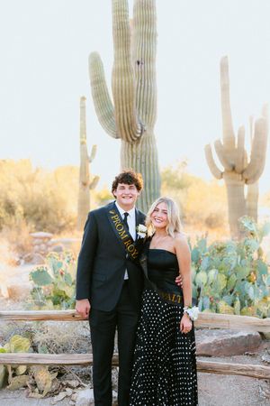 A young couple in prom attire pose before giant saguaro cacti in an Arizona desert setting.