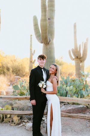 A couple in formal attire poses before tall saguaro cacti in a desert setting, she in white gown, he in black tuxedo.