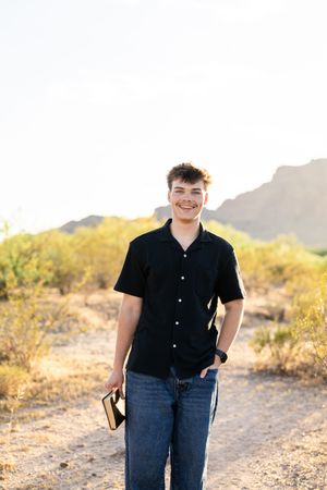 Smiling young man in black shirt holding a book on a desert trail with mountains and greenery.
