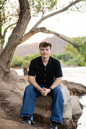 Young man in black shirt and jeans sitting on rocks by a river under a tree at golden hour.
