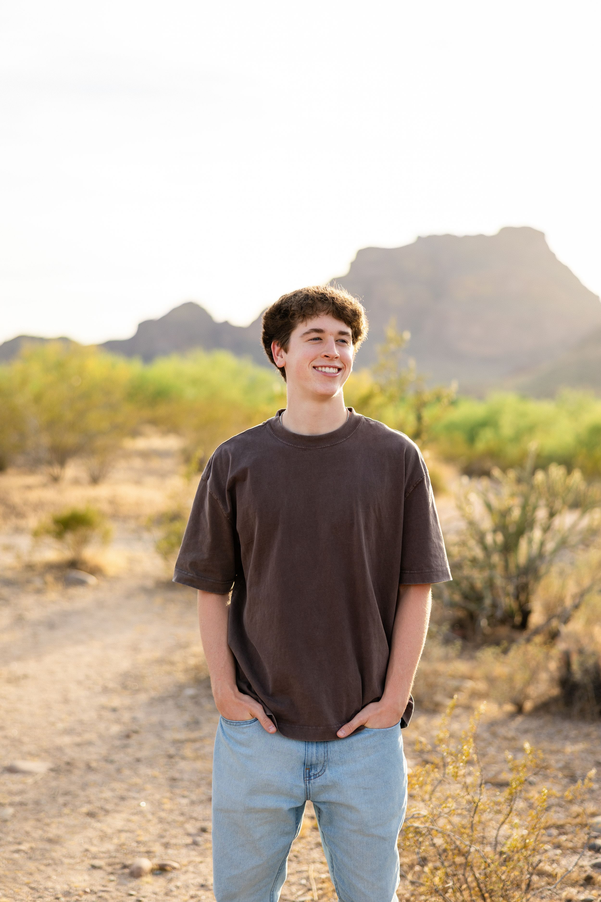 Smiling teenage boy in brown t-shirt and jeans stands on desert trail with mountain backdrop in Arizona.