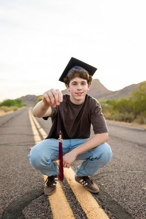 Young graduate in cap crouching on desert road holding tassel, mountains in background.