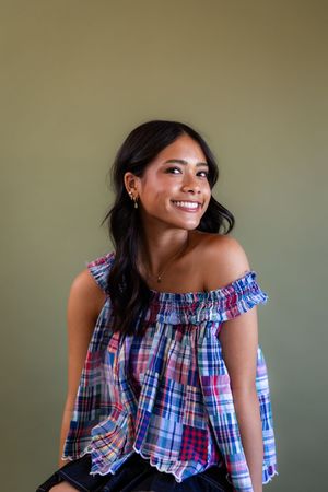 Smiling young woman in colorful plaid off-shoulder top poses against olive green background.
