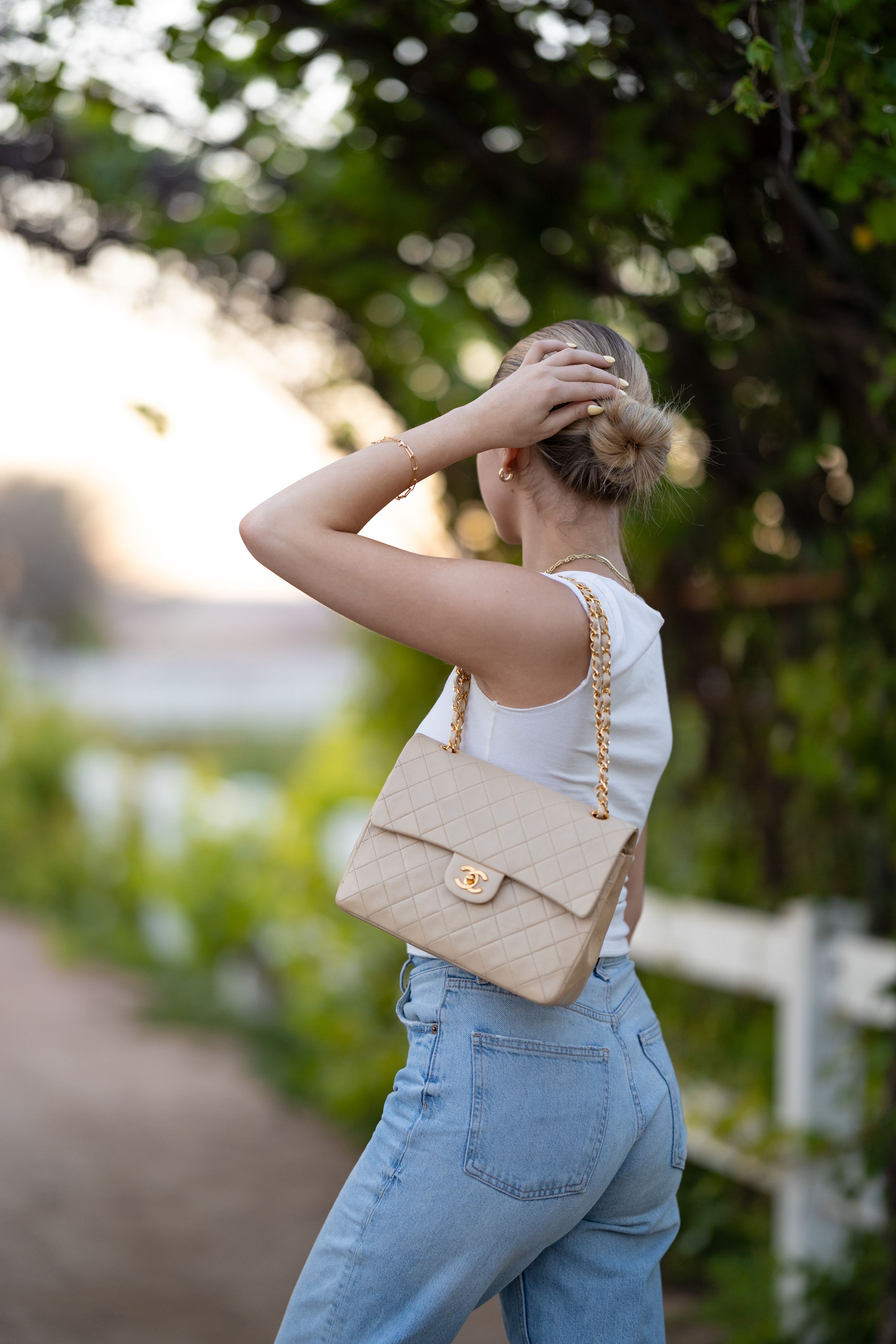Woman in white top and jeans carrying a beige quilted chain bag, standing on a garden path at golden hour.