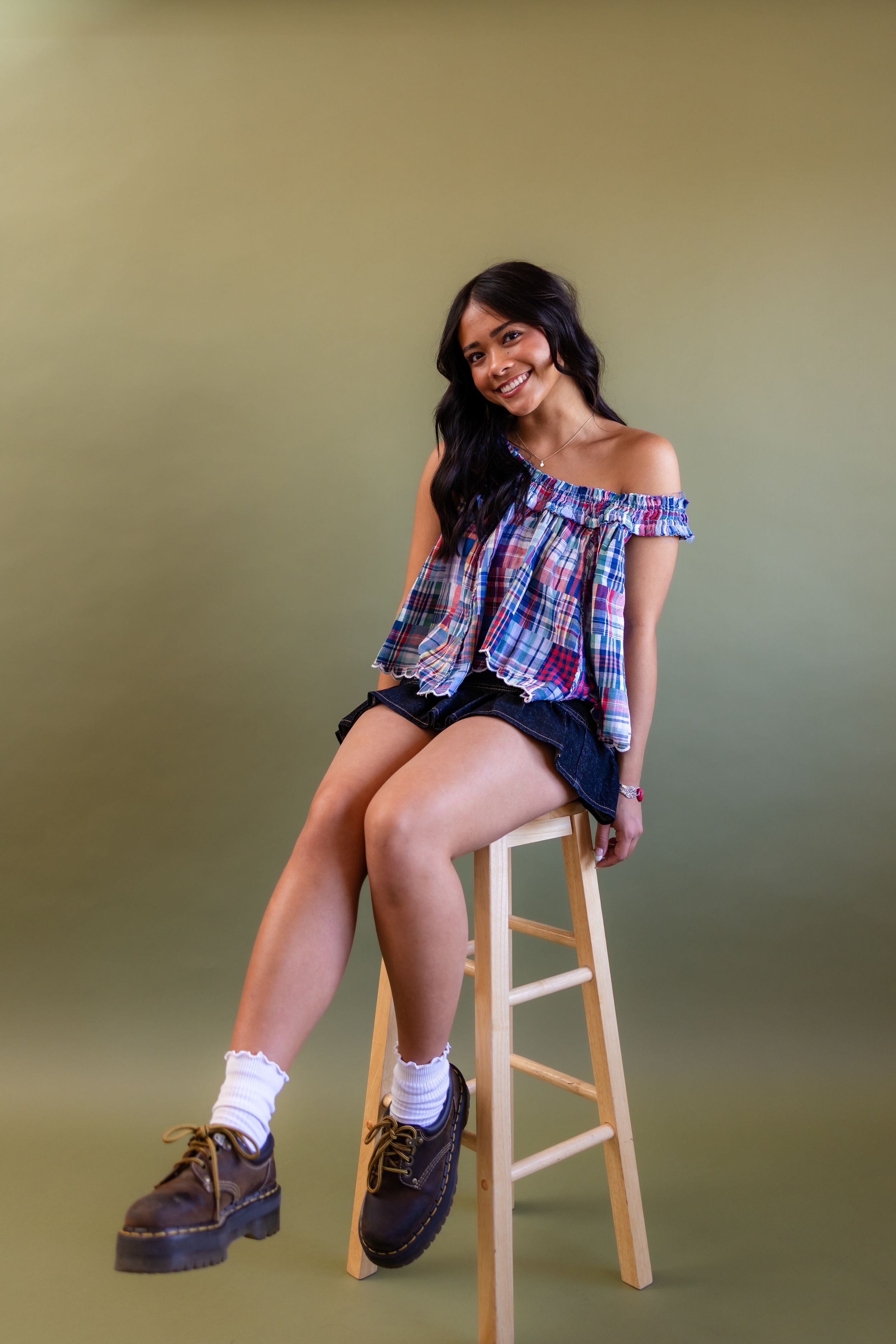 Young woman in plaid off-shoulder top, denim shorts, white socks, and brown boots sitting on a wooden stool.