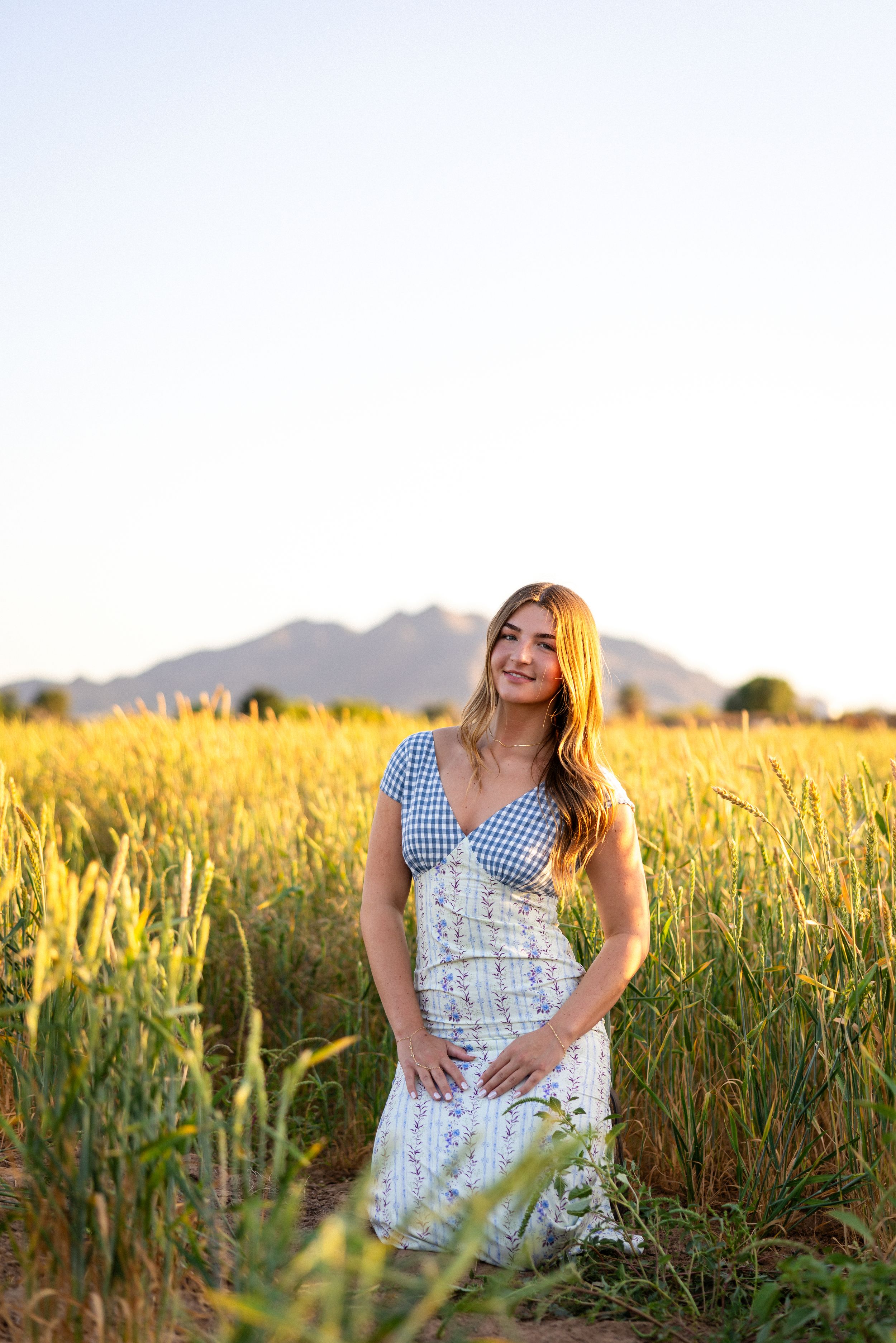 Young woman in floral dress standing in golden wheat field with mountain backdrop at sunset.