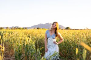 A young woman in a floral dress stands in a golden wheat field with mountains in the background at sunset.