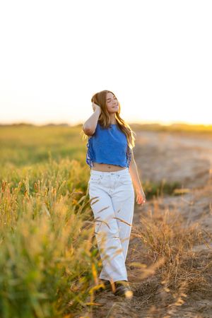 Young woman in blue crop top and white wide-leg jeans walking through golden wheat field at sunset.