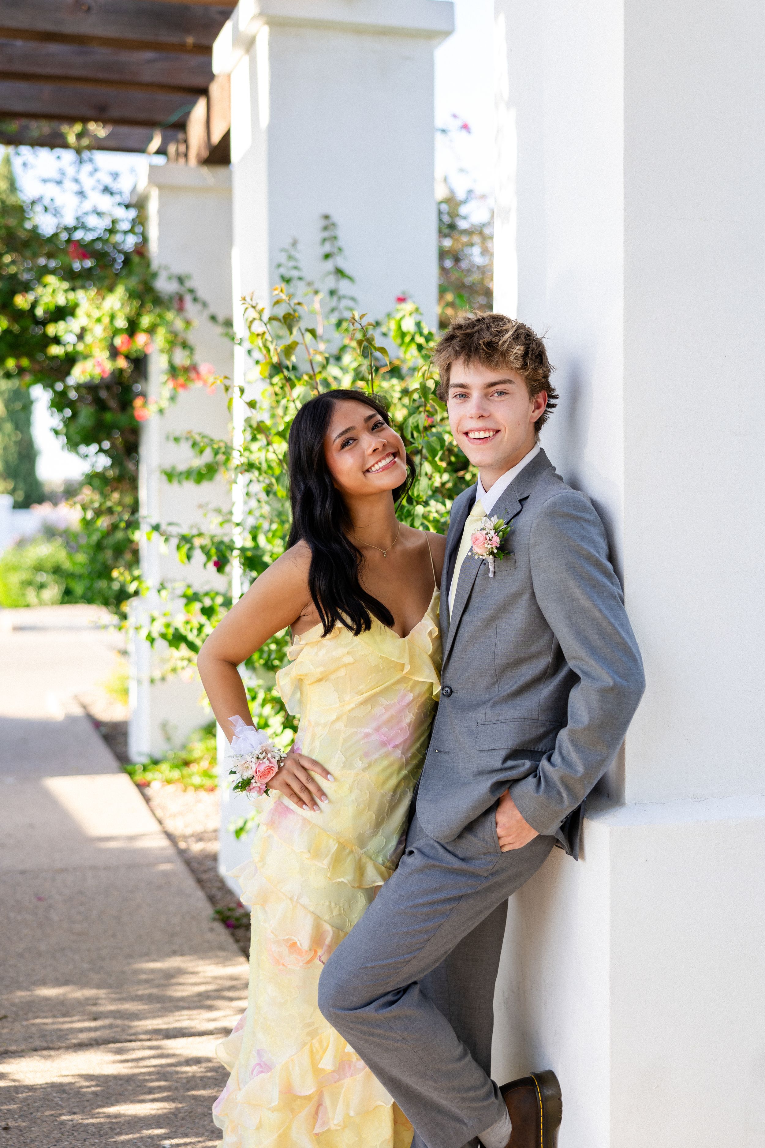 A young couple dressed in prom attire, posing outdoors by white columns with lush greenery in the background.
