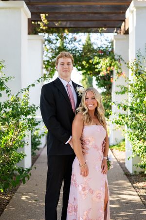 A young couple dressed in formal prom attire poses under a pergola surrounded by lush greenery.