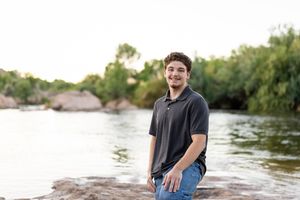 Young man smiling by a river, standing on rocks with lush green trees in the background on a sunny day.