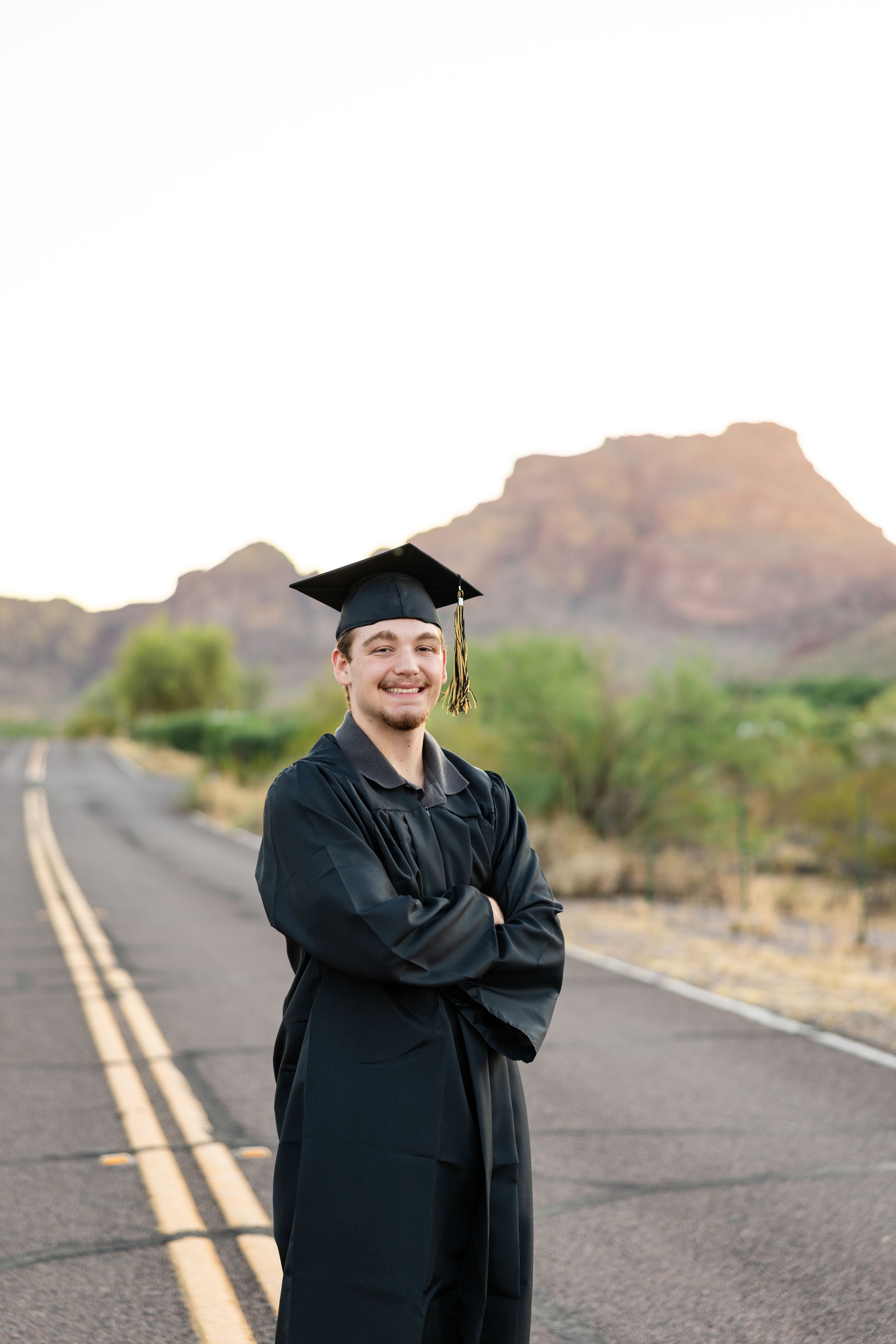 Smiling graduate in black cap and gown stands on desert road with red rock mountains in background.