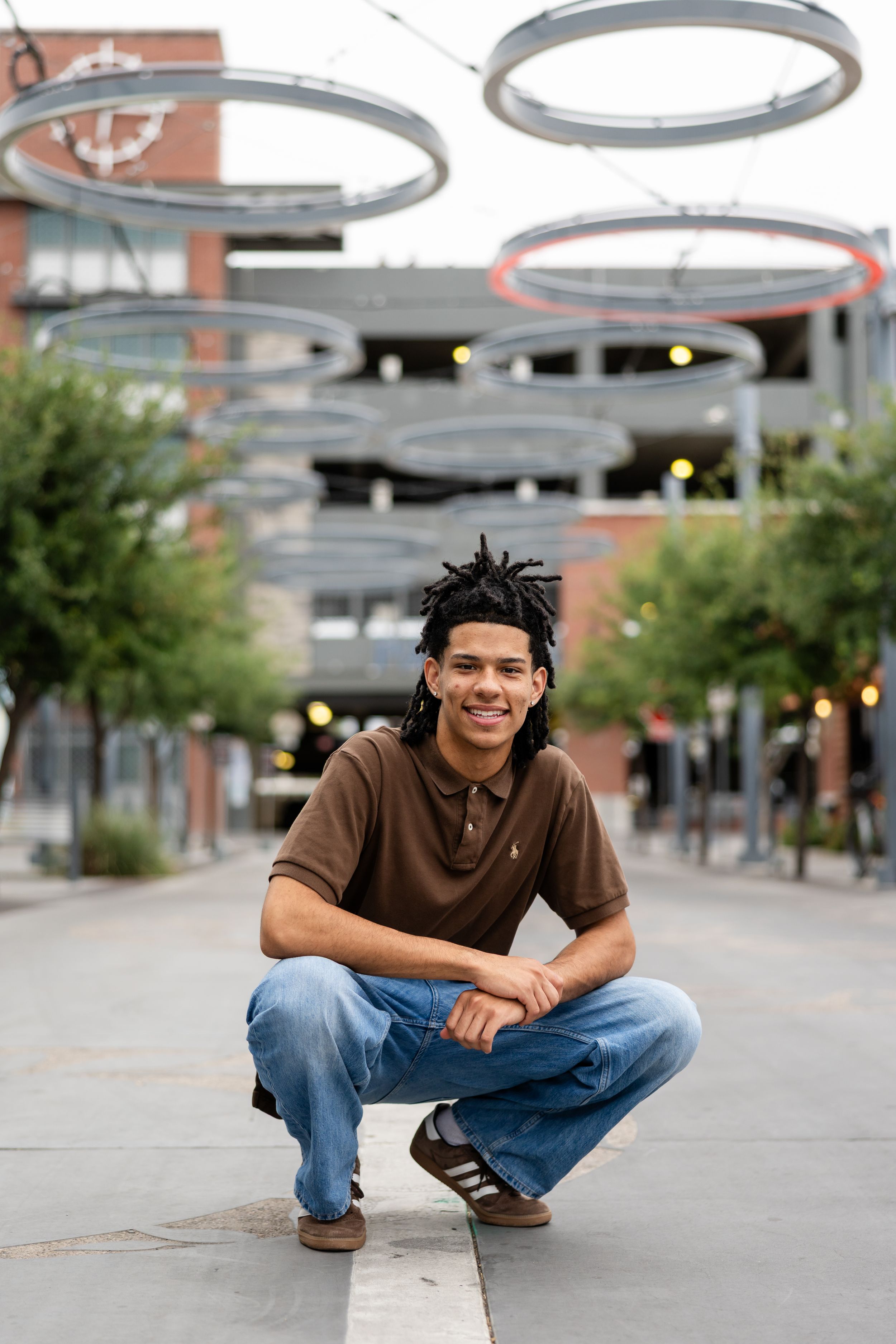 Young man with dreadlocks crouching and smiling in an urban plaza with circular light installations overhead.