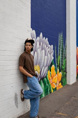 Young man in brown shirt and jeans leans against white brick wall with colorful floral mural backdrop.