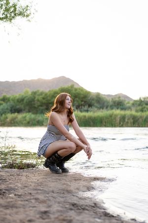 A smiling redhead woman crouches by a river in a striped dress and boots, with mountains and greenery in the background.