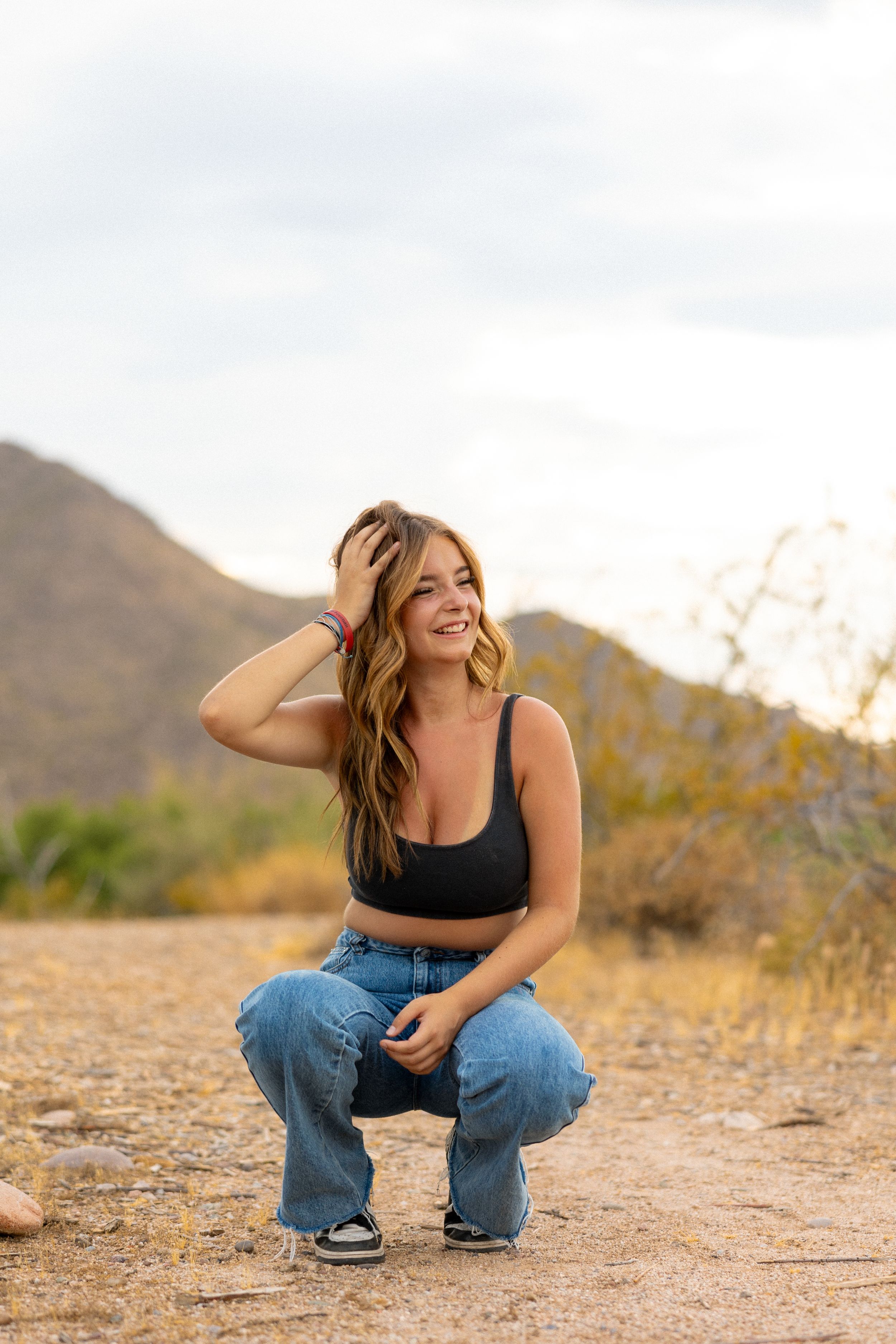 Young woman in black crop top and jeans crouching outdoors with desert mountains in background, smiling and touching her hair.
