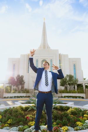 Young man in blue suit poses triumphantly before a white LDS temple spire amid colorful gardens on a sunny day.