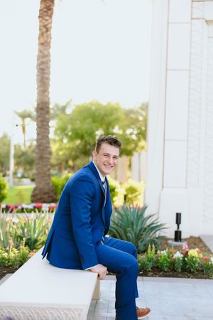 Young man in a blue suit sitting outdoors near a white building with palm trees and colorful flowers in the background.
