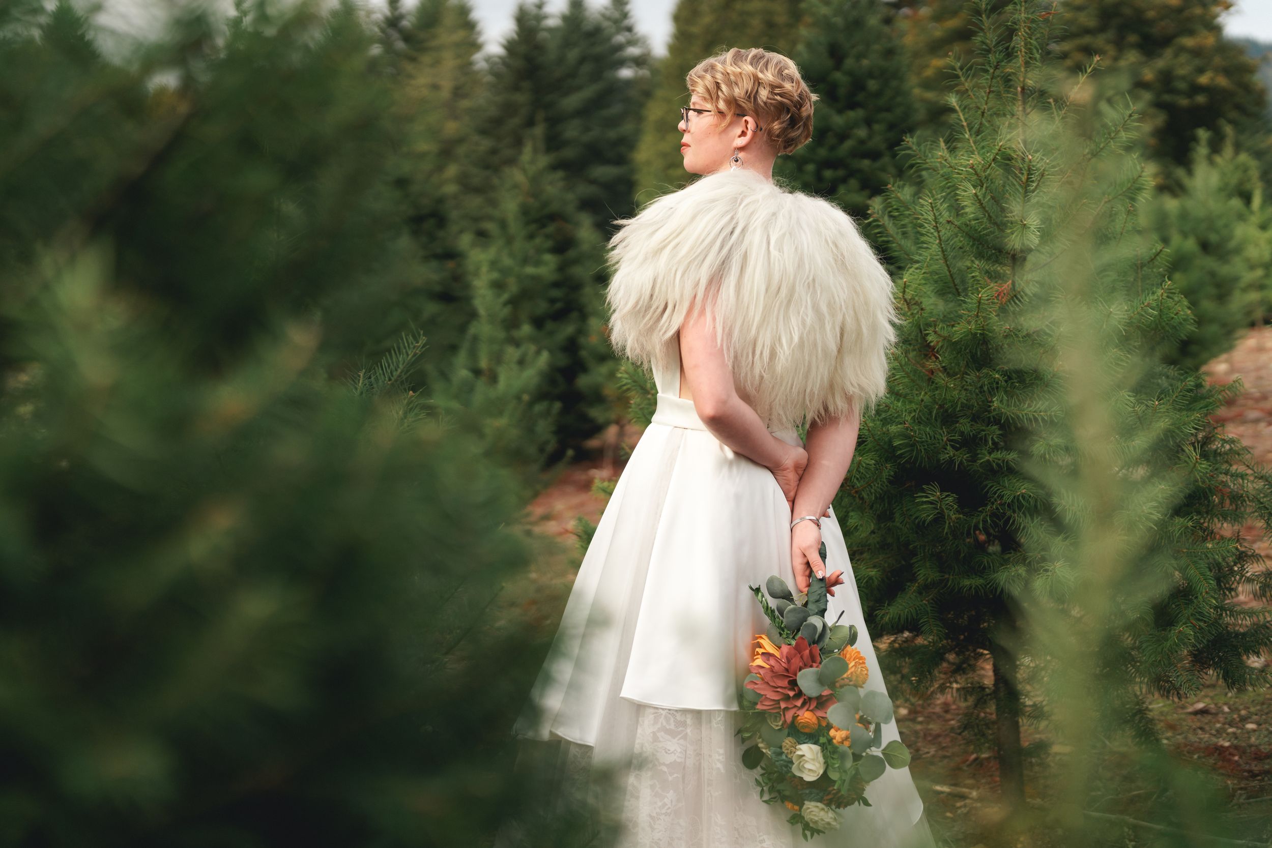 A bride in a white wedding dress walks through a lush green forest setting with sunlight filtering through the trees.