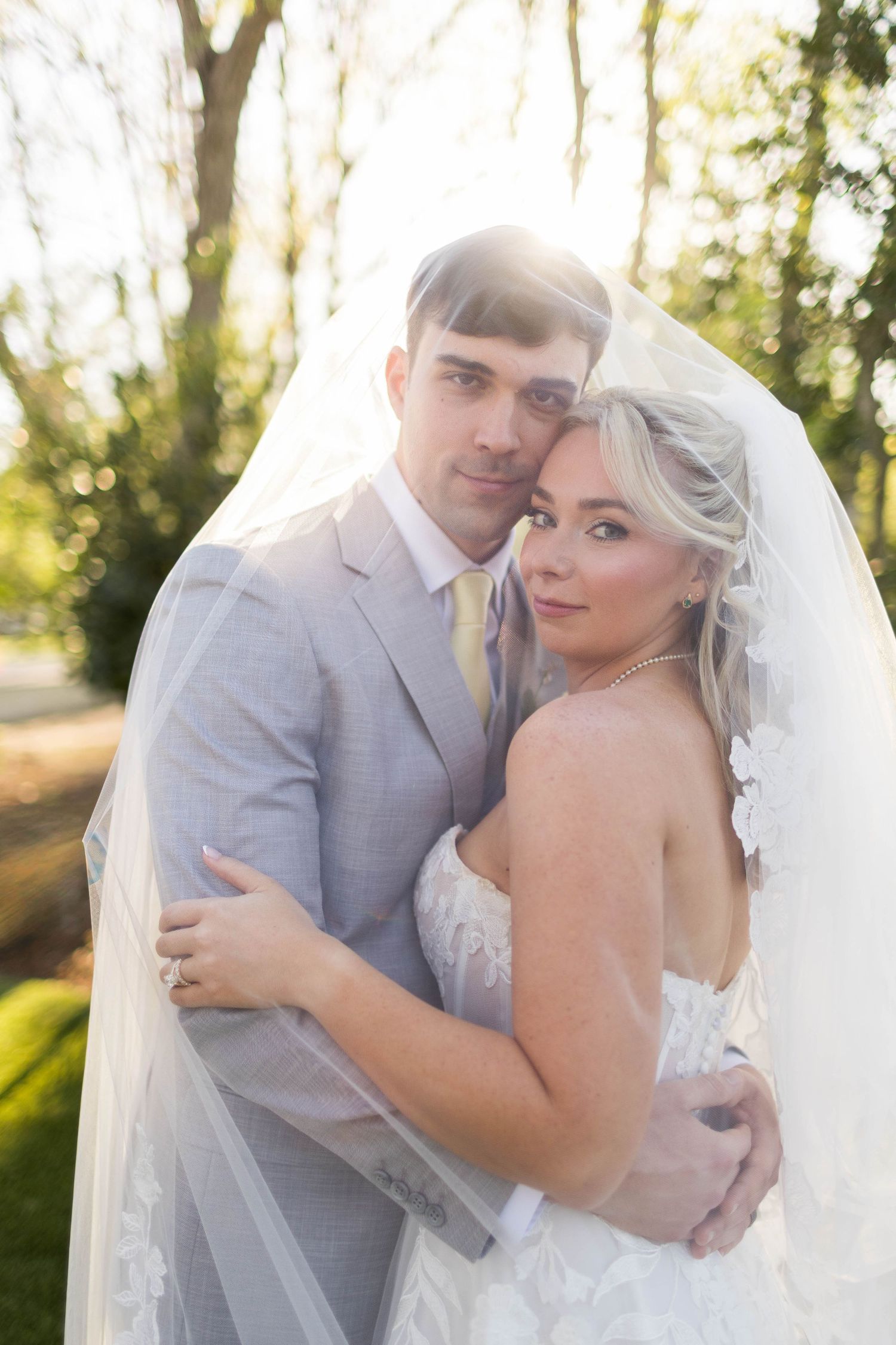 Groom embraces smiling bride from behind at golden hour, bride holding a pastel floral bouquet.