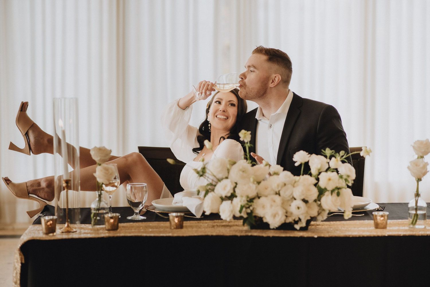 A couple at their wedding reception head table sharing a lighthearted moment with white floral centerpieces.